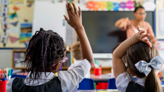 Pupils in a classroom. One pupil has their hand up trying to get the attention of the teacher at the front of the classroom 