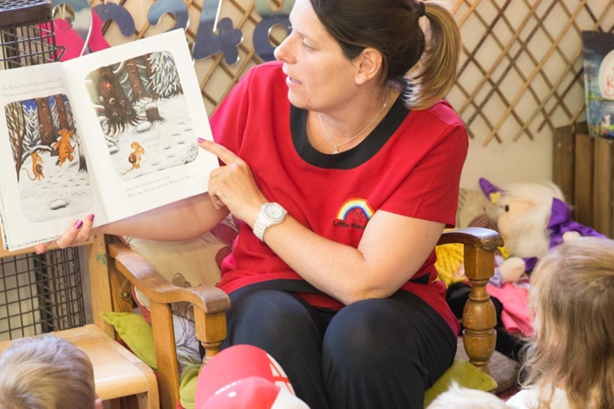 Teacher holding up a story book and reading to a class of small children.