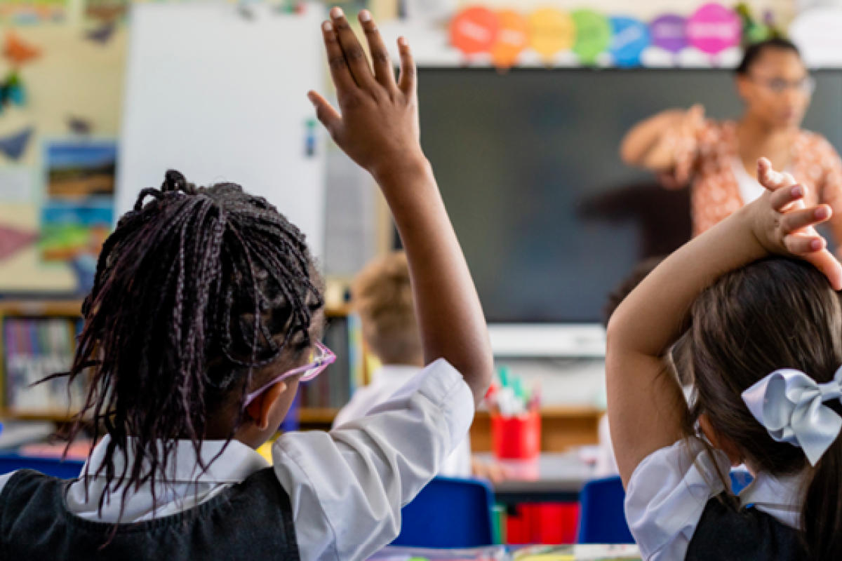 Pupils in a classroom. One pupil has their hand up trying to get the attention of the teacher at the front of the classroom 